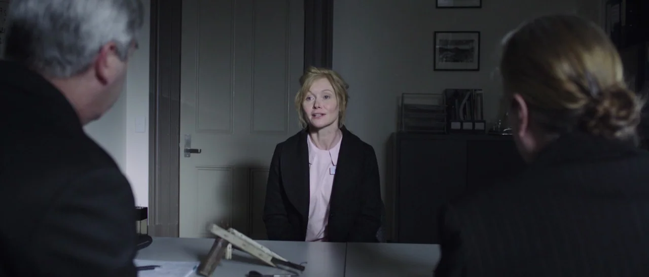The Babadook (2014) movie still: medium — A dimly lit room with a grey table and two people facing a woman. The woman sits at the table while the…