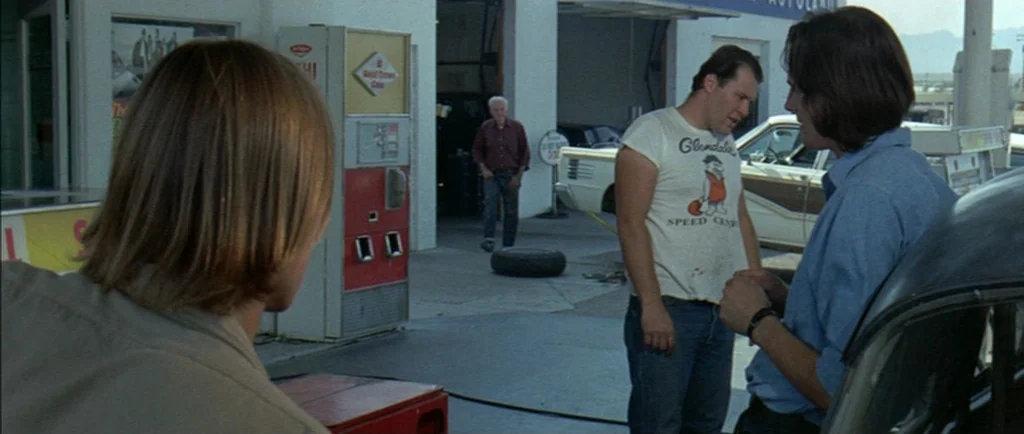 Two Lane Blacktop (1971) movie still: medium — A sunlit automotive service area features a soda vending machine and concrete pavement. Three men…