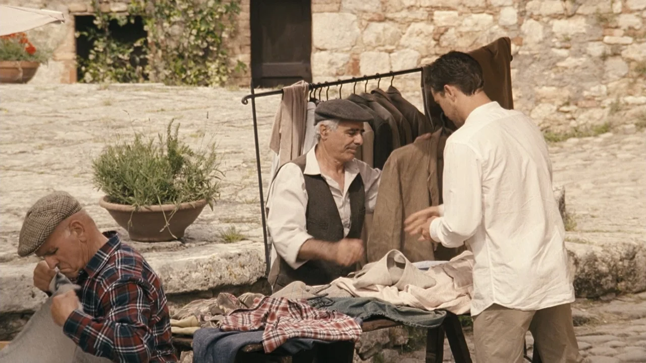 Shadows in the Sun (2005) movie still: medium — An outdoor market stall features an elderly man helping a customer select garments. The setting…