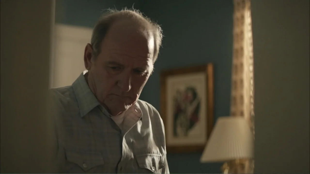 The Hollars (2016) movie still: medium — A quiet indoor space features dark blue walls adorned with a framed picture. An elderly man in a plaid…