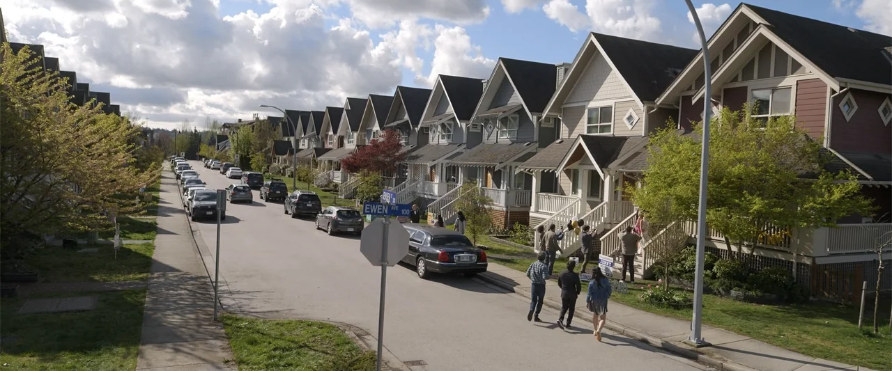 Final Destination Bloodlines (2025) movie still: wide — A quiet residential street lined with suburban houses and parked cars under a bright sky. A…