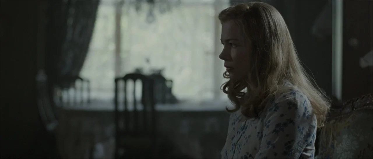 Suite Française (2015) movie still: medium — A dimly lit room with an out of focus dining table and chairs near a window. A woman sits in the…