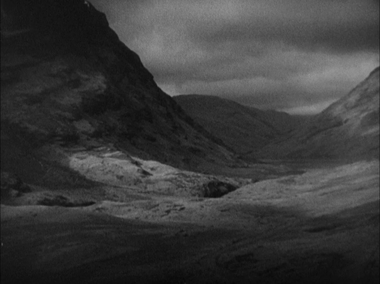 The 39 Steps (1935) movie still: establishing — A desolate mountain valley is framed by steep, rocky slopes under a heavy, clouded sky. Patches of…