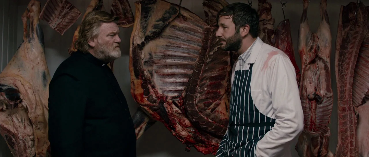 Calvary (2014) movie still: medium — A cold storage room with several large animal carcasses hanging from metal hooks. A priest and a butcher stand…