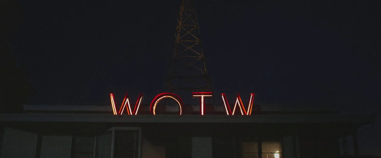 The Vast of Night (2019) movie still: wide — A dark night exterior features a glowing red neon sign reading WOTW. A metal broadcast tower stands…