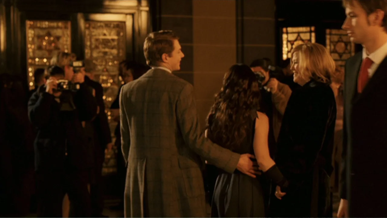 Penelope (2007) movie still: medium — A dimly lit lobby features ornate, patterned glass doors in the background. A man standing behind a woman…