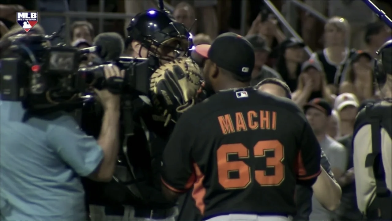 Ferrell Takes the Field (2015) movie still: medium — A professional baseball player wearing a jersey stands near a masked catcher in a dugout. A…