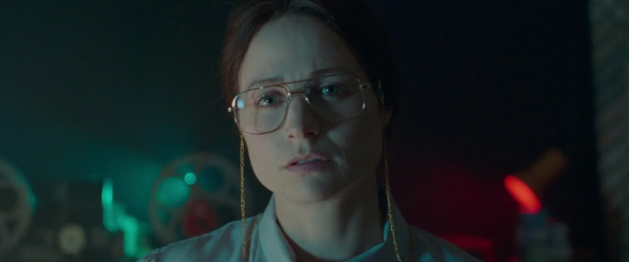 Censor (2021) movie still: closeup — A dark, cluttered archive room contains film reels and technical equipment in the background. A woman wearing…