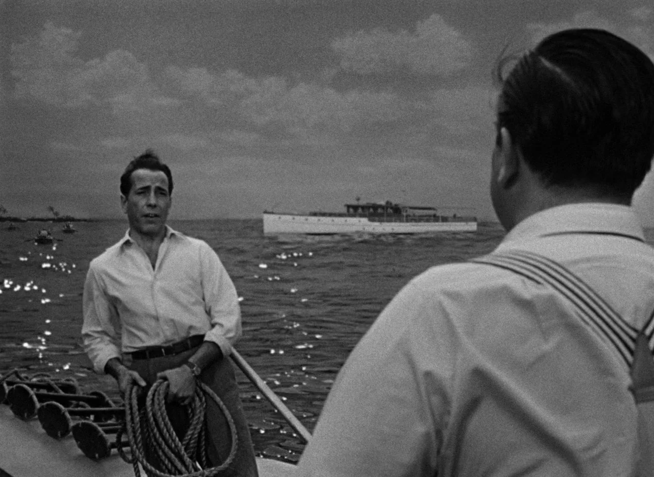 Key Largo (1948) movie still: medium — A man stands on the deck of a boat with a yacht visible at sea. He holds a coiled length of rope while…