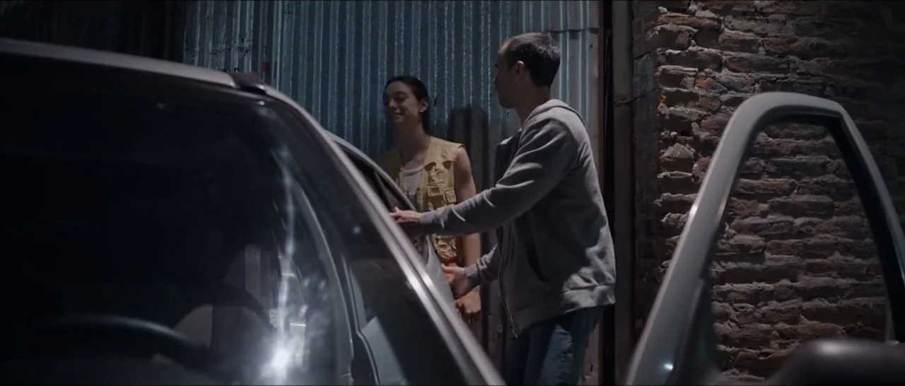 Gunman (2025) movie still: medium — An exterior alleyway with corrugated metal walls and a brick pillar at night. A man in a hoodie opens a car door…