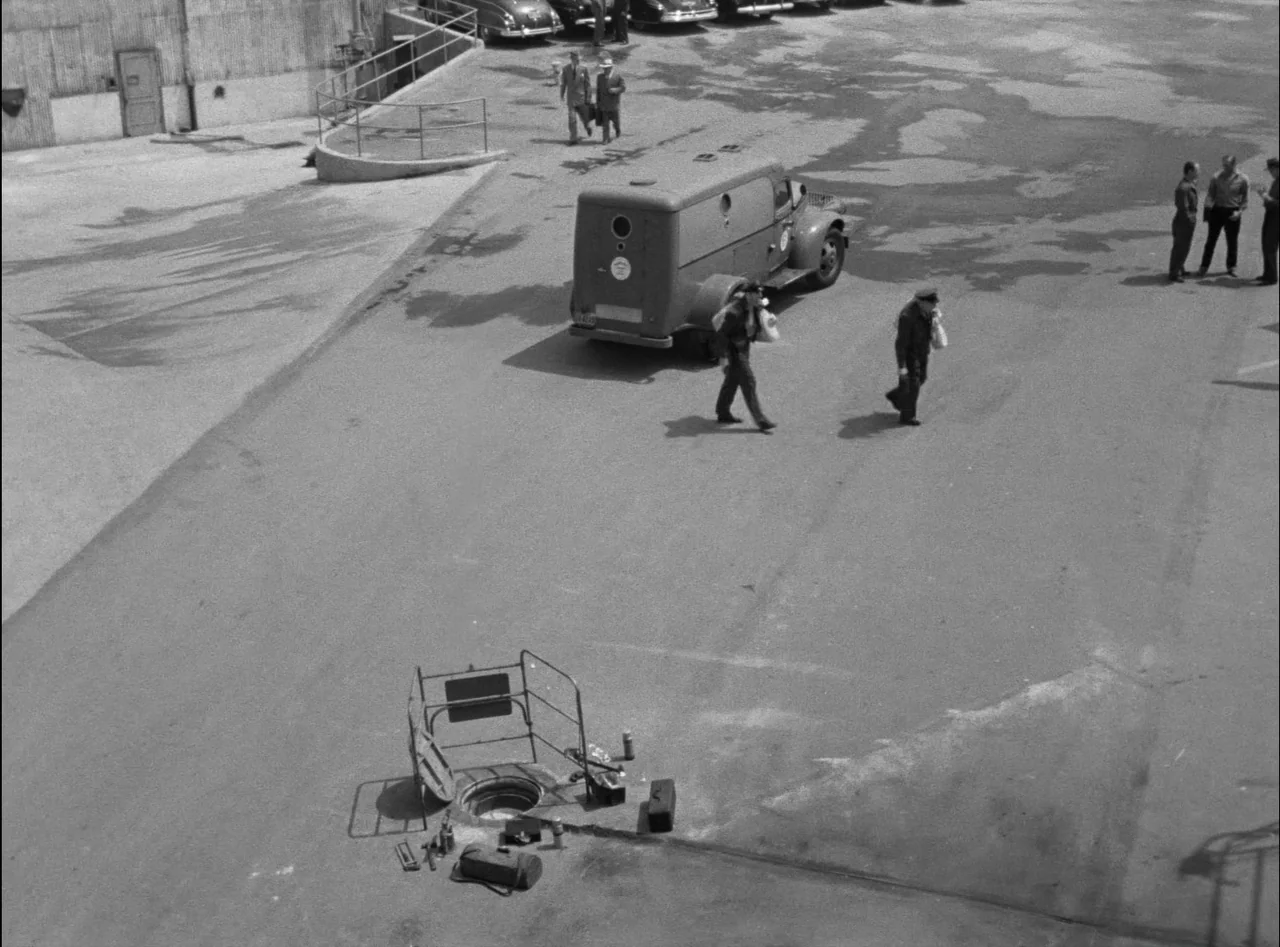 Criss Cross (1949) movie still: wide — An industrial paved area features an open manhole surrounded by metal safety railings. Several men walk…