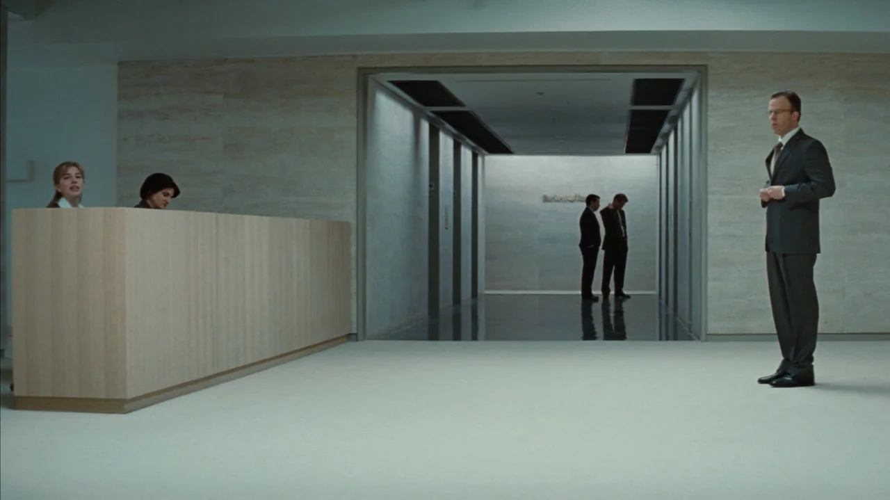 Duplicity (2009) movie still: fullbody — A minimalist corporate building entrance features light wood paneling and stone flooring. A man in a suit…