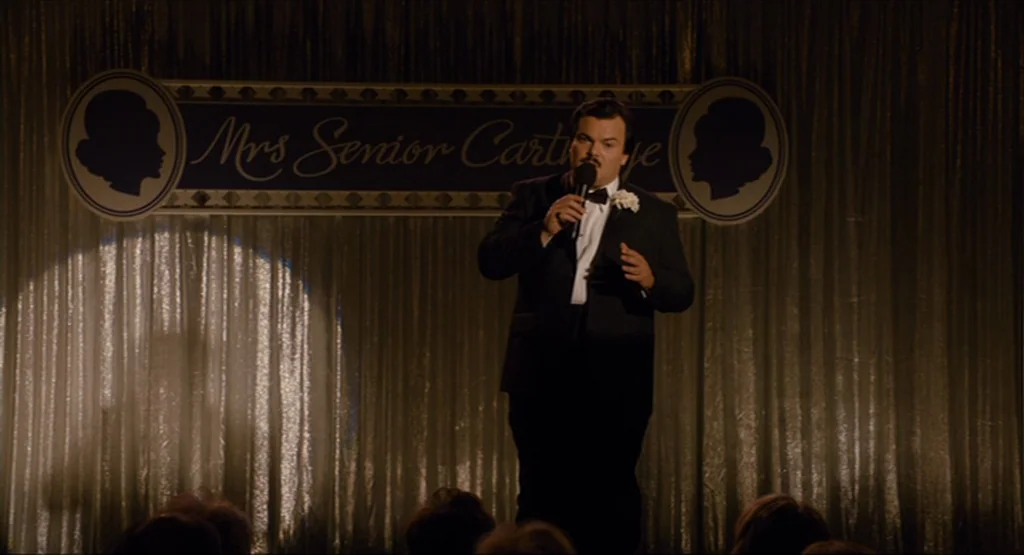 Bernie (2011) movie still: fullbody — A dimly lit stage features a gold shimmering curtain backdrop with a banner above. A man in a formal tuxedo…