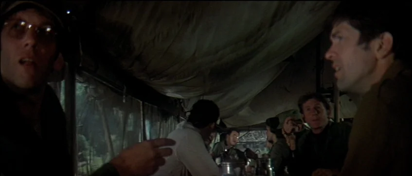 MASH (1970) movie still: medium — The interior of a canvas-walled mess tent features men seated around a table. A man on the left talks while…