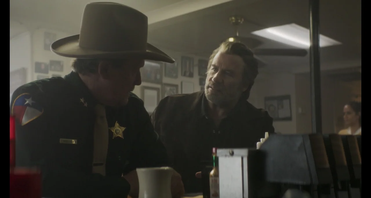The Poison Rose (2019) movie still: medium — A diner booth with framed photographs on the wall and a dark table. A sheriff in uniform talks to a man…