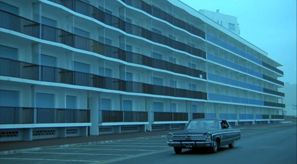 Un Flic (1972) movie still: wide — A long, multi-story modern apartment building with blue balcony panels stretches across the frame. A dark vintage…