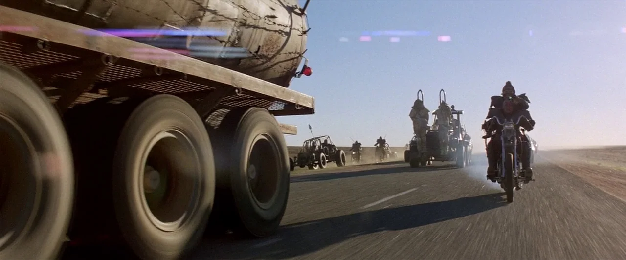 Mad Max 2 (1981) movie still: wide — A long, flat asphalt highway runs through a barren desert landscape under a clear sky. A convoy of armored…