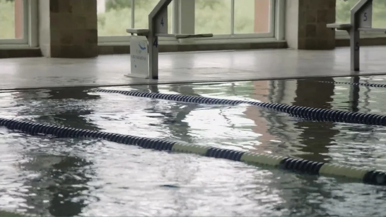Dear Rider: The Jake Burton Story (2021) movie still: establishing — An indoor swimming pool features numbered starting blocks along a tiled deck…