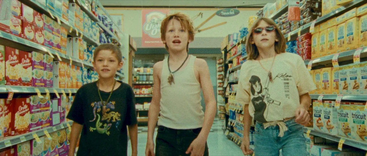 Riddle of Fire (2023) movie still: fullbody — A well-lit grocery store aisle packed with colorful snack boxes on metal shelves. Three young children…