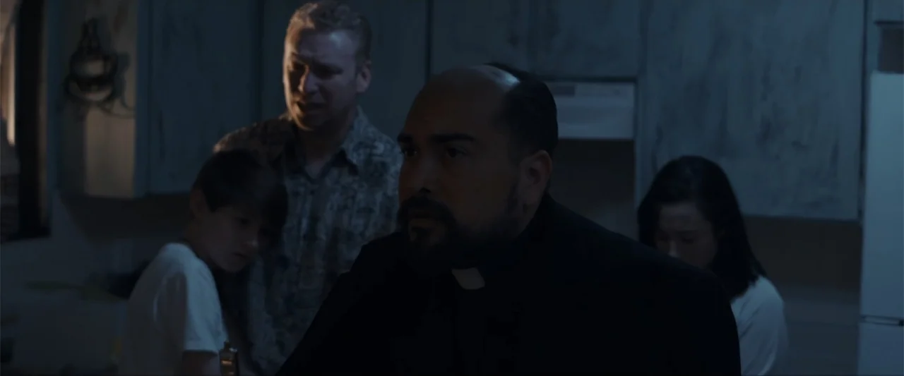 Anomaly (2006) movie still: medium — A dimly lit room with muted cabinetry and dark shadows surrounding the subjects. A priest and two men stand…