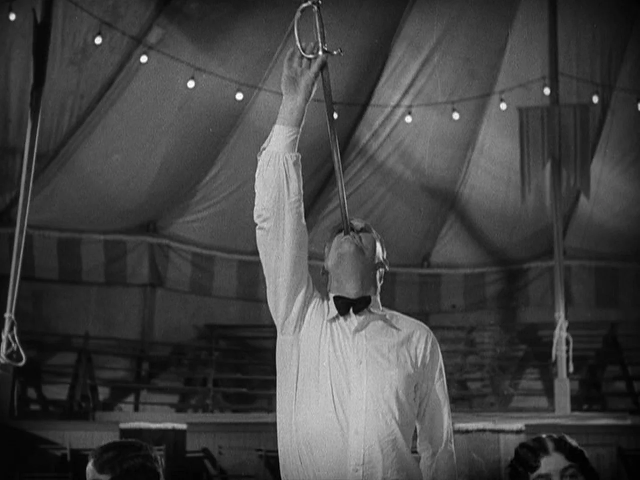 Freaks (1932) movie still: medium — A man in a circus tent performs a sword-swallowing act for an audience. He is positioned in the center, tilting…