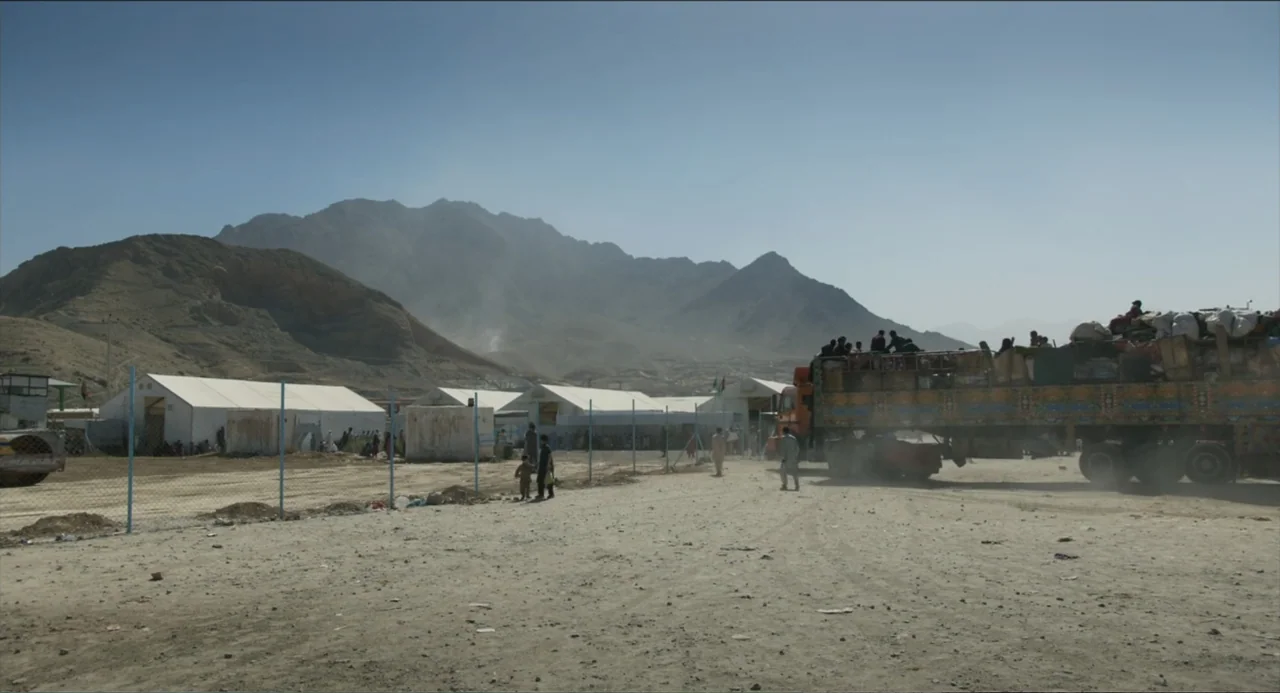 Human Flow (2017) movie still: wide — A dusty encampment features rows of white tents under a mountain range. A large cargo truck carrying many…