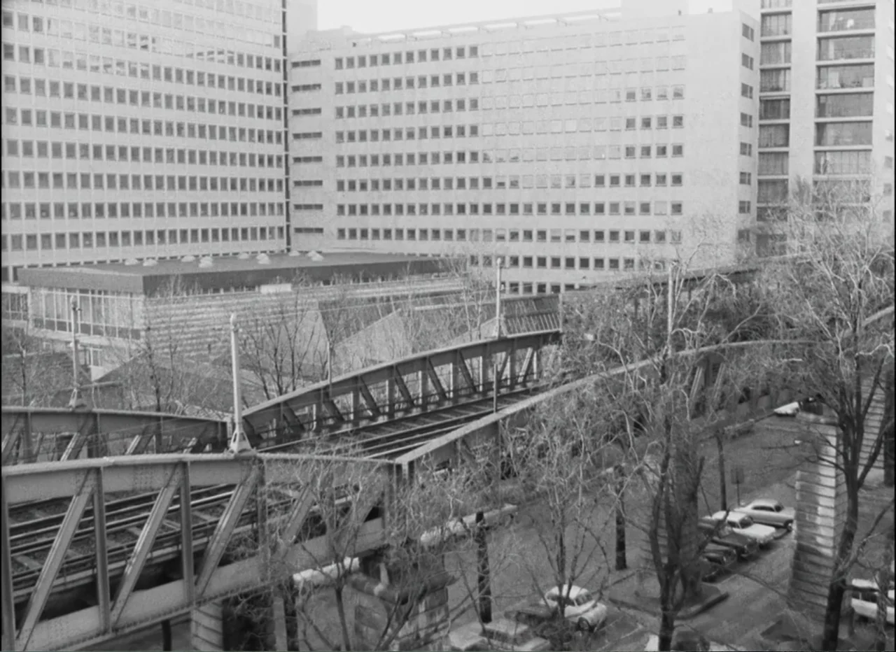 Masculin Féminin (1966) movie still: wide — An elevated train track cuts across an urban landscape with large modernist apartment buildings. Parked…