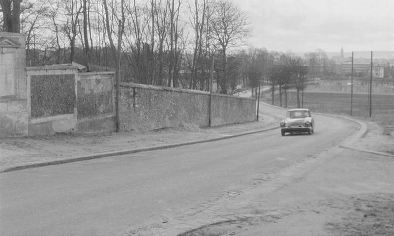 Eyes Without A Face (1960) movie still: wide — An open asphalt road winds past a long, weathered stone wall and bare trees. A small vintage European…