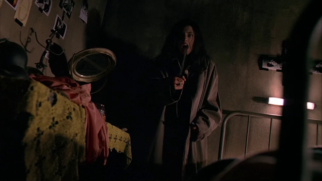 Ginger Snaps (2000) movie still: medium — A dimly lit room contains a bed with yellow bedding and wall photos. A woman in a trench coat stares…