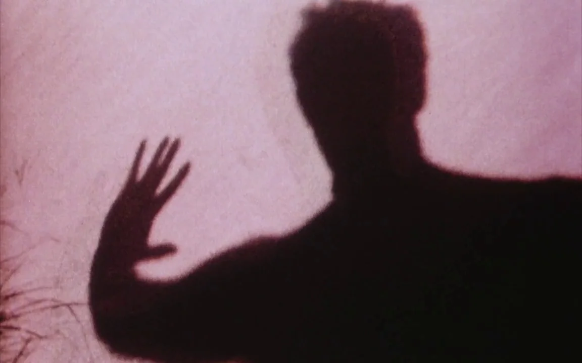 The Last of England (1987) movie still: closeup — A dark human shadow is cast onto a flat textured surface. The shadow figure extends one arm with…