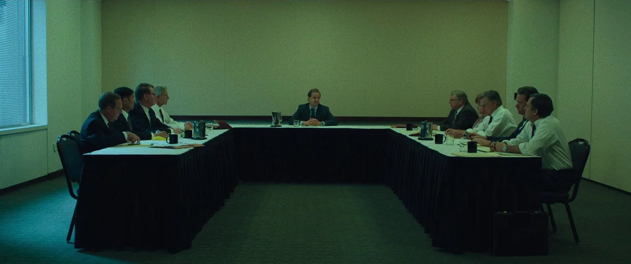 Dark Waters (2019) movie still: wide — A windowless corporate meeting room features a large U-shaped table arrangement with twelve attendees. The…