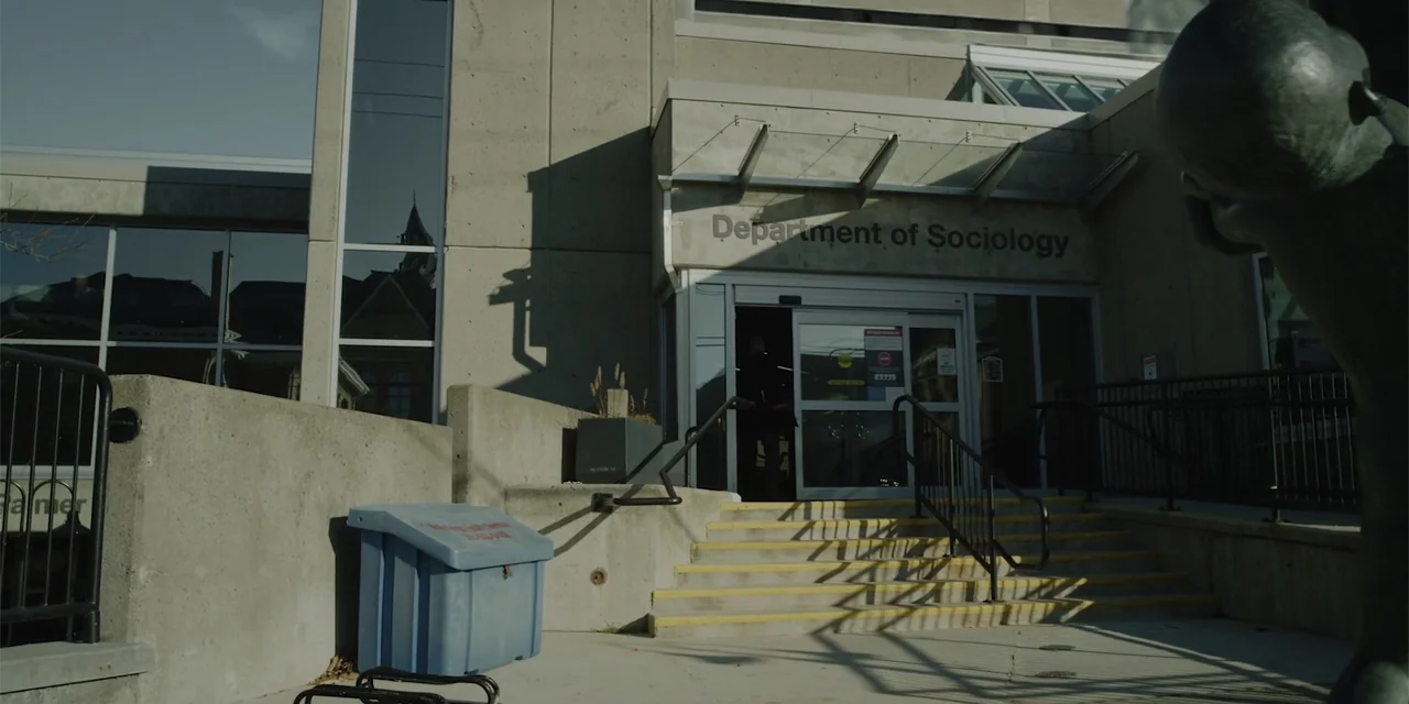 Lobos (2019) movie still: wide — A modern concrete building entrance labeled Department of Sociology sits in bright daylight. The building features…