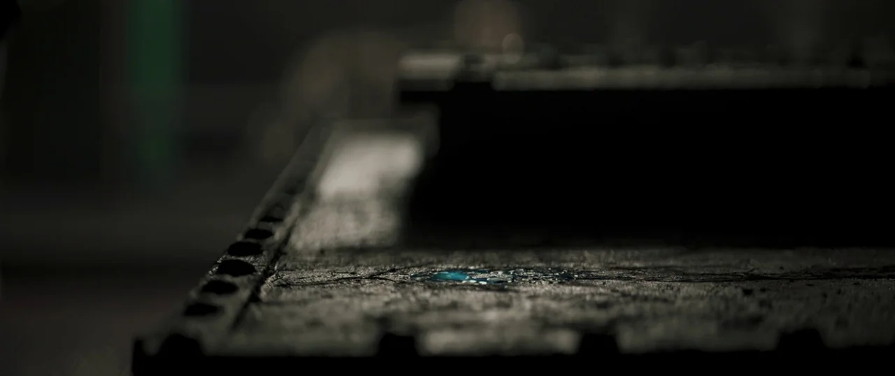 Sea Fever (2019) movie still: closeup — A dark, textured metal surface sits in a dim, shadowed industrial area. Small pools of blue liquid glisten…