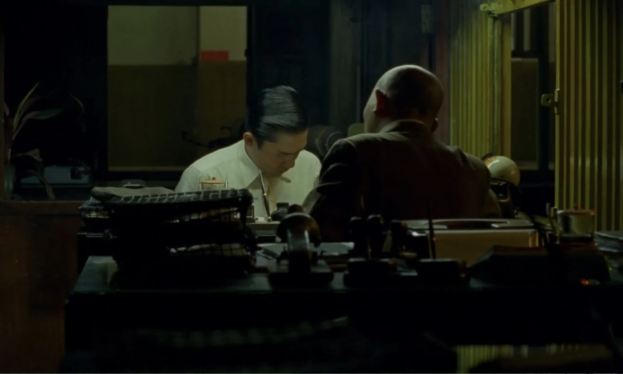 In the Mood for Love (2000) movie still: medium — A cluttered 1960s office is illuminated by a single warm desk lamp at night. Two men are seated at…
