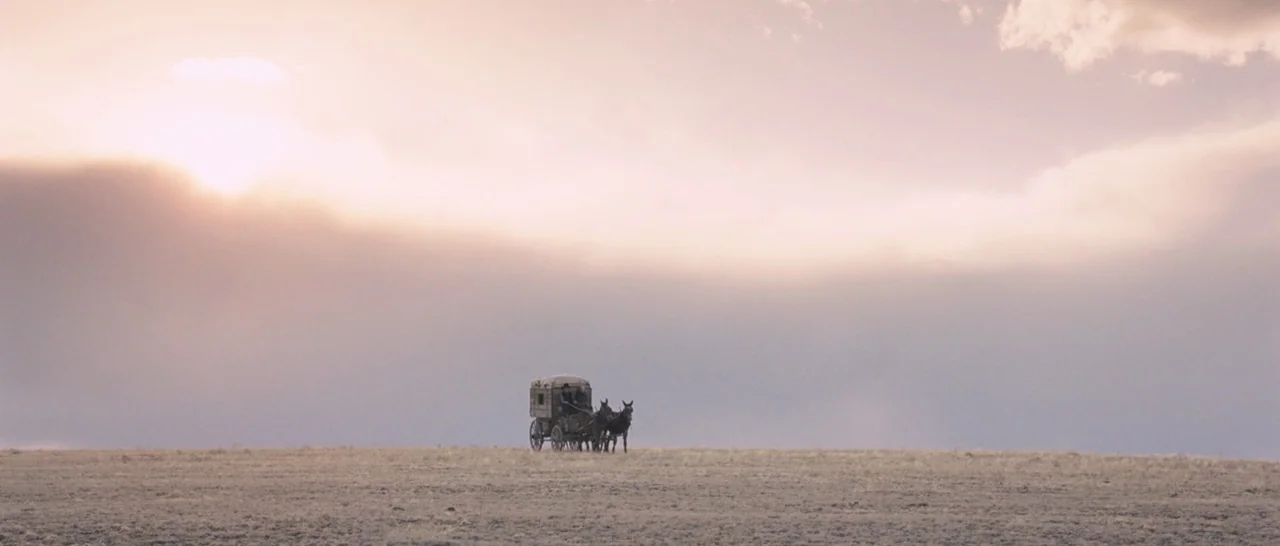 The Homesman (2014) movie still: wide — An open expanse of dry grass stretches toward a vast, hazy, light-filled sky. A small carriage pulled by a…