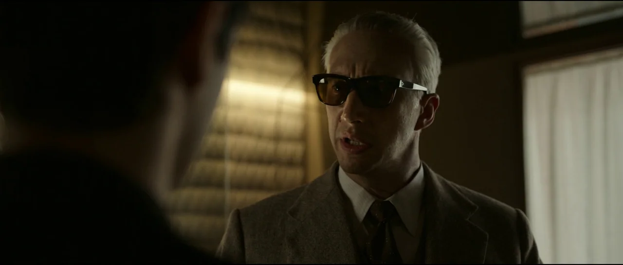 Ferrari (2023) movie still: medium — A dimly lit room with textured, paneled walls and a blurred window frame visible. A man with slicked-back white…
