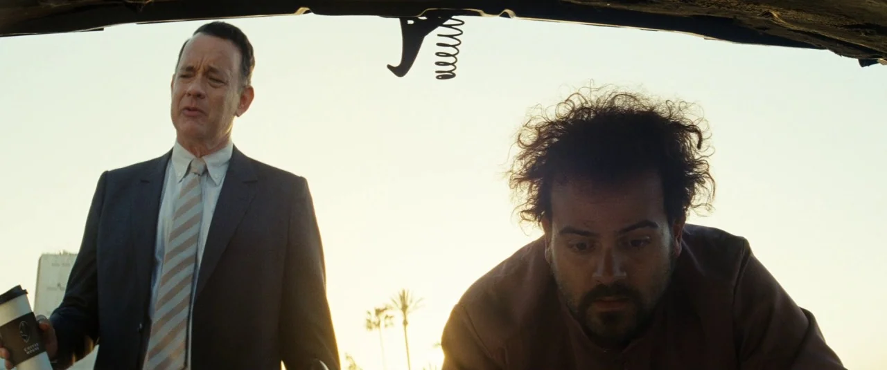A Hologram for the King movie still: medium — Two men stand near an open vehicle trunk under the bright afternoon sun. The older man in a suit looks…