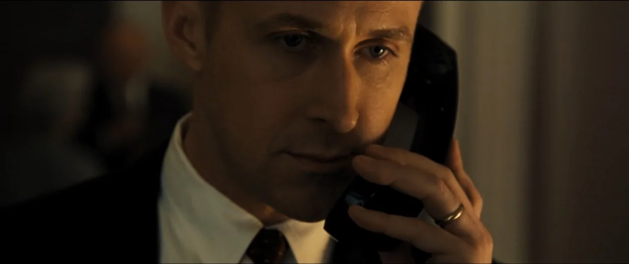 First Man (2018) movie still: closeup — A dark, minimally lit room with indistinct architectural details and muted textures. A man in a dress shirt…