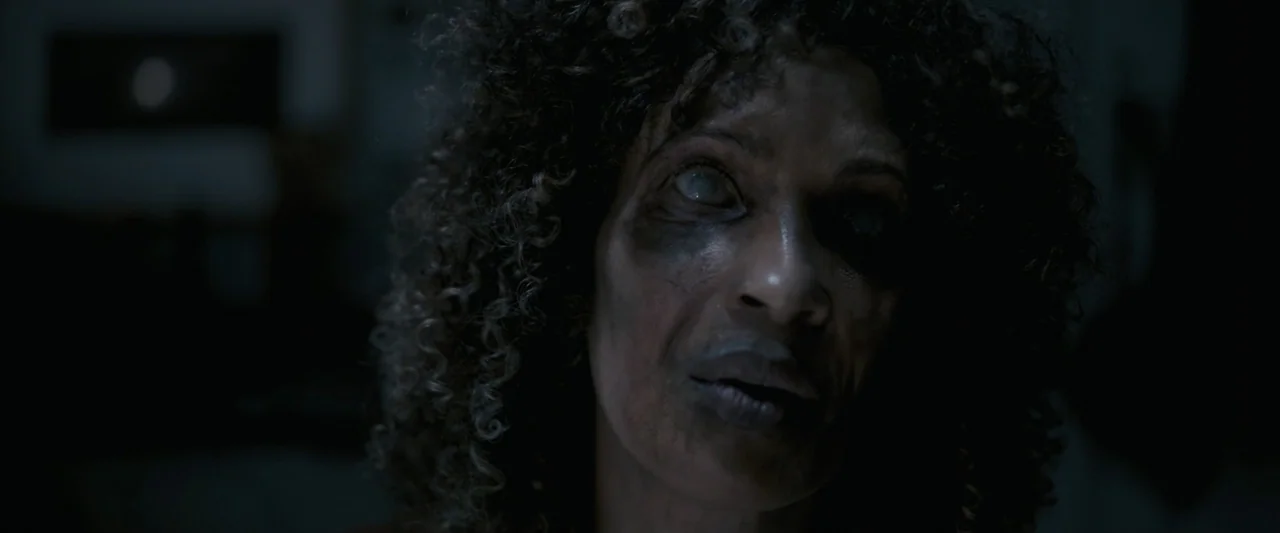 Talk to Me (2023) movie still: closeup — A dimly lit indoor space with shadows obscuring the background. A woman with textured curly hair stares…