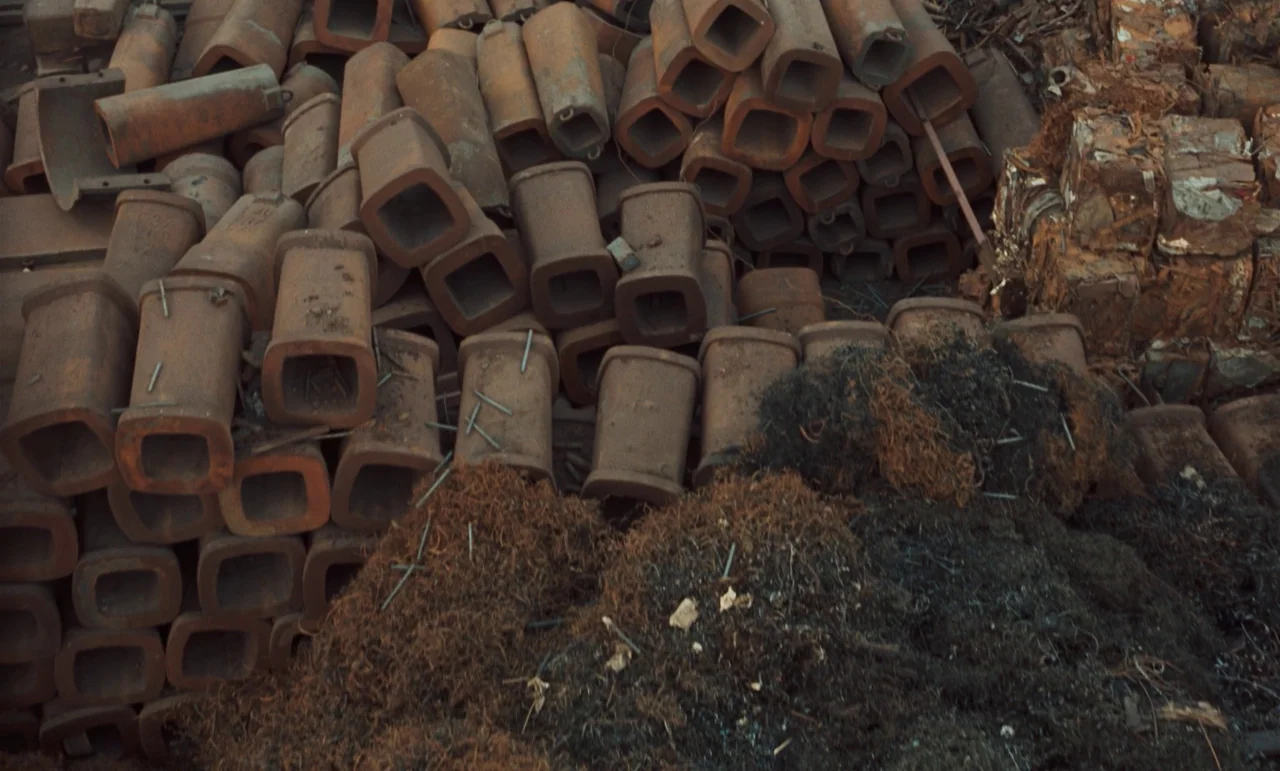 Larks On A String (1969) movie still: wide — A collection of hollow rectangular metal pipes are stacked in a large weathered pile. Mounds of dark…