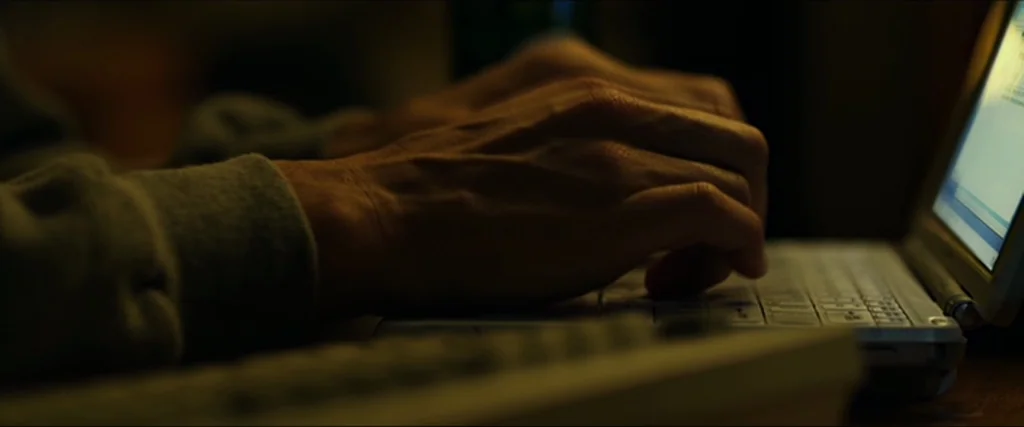 The Social Network movie still: closeup — A dark environment centered on the glowing keys of a portable laptop. A pair of hands rests on the…