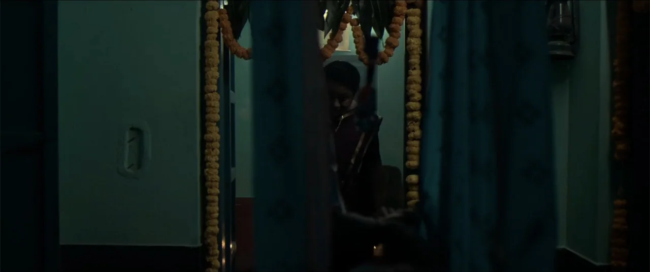 Cheekatilo (2026) movie still: medium — A dimly lit residential threshold framed by marigold garlands and heavy blue curtains. A woman in a dark…