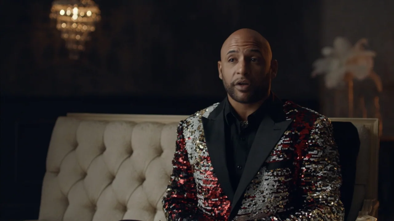 The Stroll (2023) movie still: medium — A dimly lit interior space features a man seated on a cream tufted sofa. A blurred crystal chandelier is…