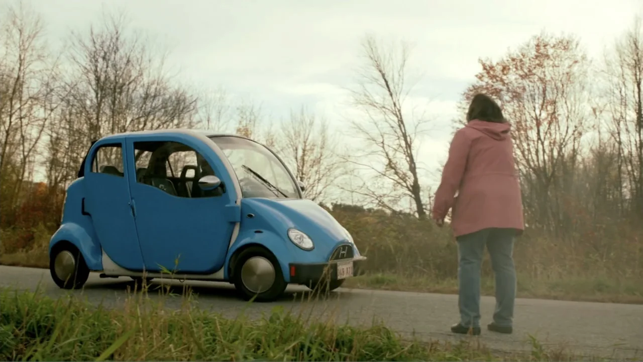 Clear History (2013) movie still: medium — A woman stands on a rural asphalt road next to a small blue electric car. Bare autumn trees line the…