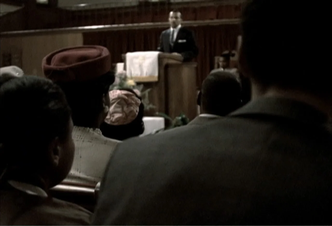 Boycott (2001) movie still: medium — A church interior with dark wooden pews and a man standing at a podium. The congregation listens while facing…