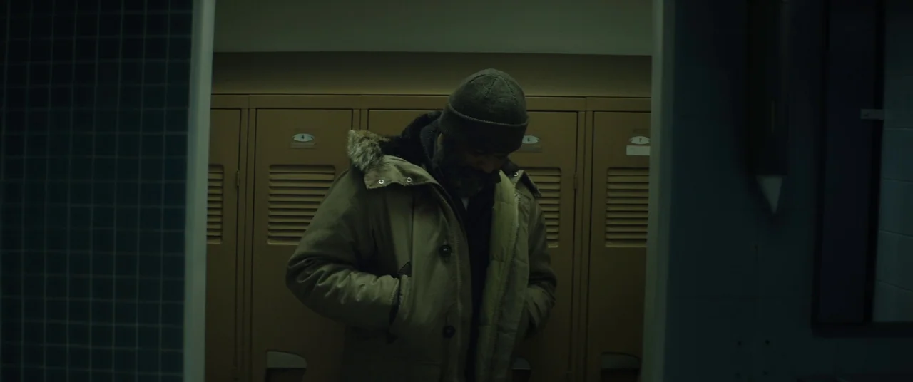 Hold The Dark (2018) movie still: medium — A locker room with rows of numbered tan metal lockers against a plain wall. A man in a parka and beanie…