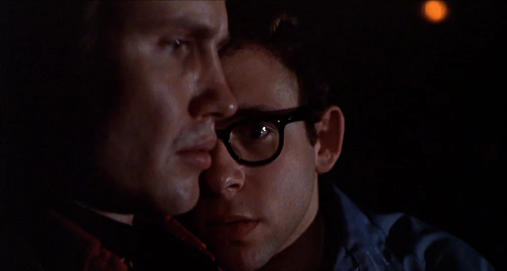 Midnight Cowboy (1969) movie still: closeup — Two men are positioned closely together in a dimly lit, nondescript dark space. The individuals appear…