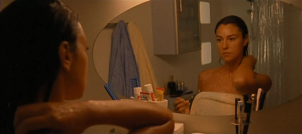 Irréversible (2002) movie still: medium — A dimly lit bathroom features a vanity mirror and various toiletries in a holder. The woman stands before…