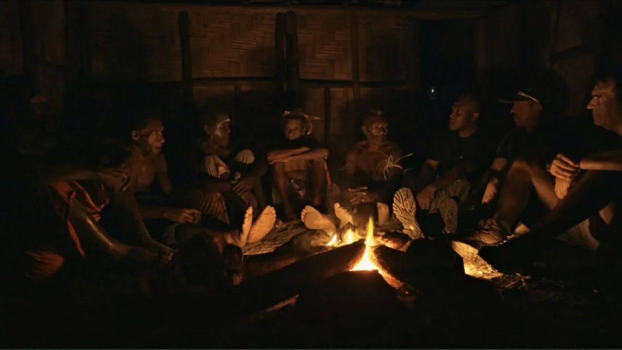 Finding Amelia (2024) movie still: medium — A dimly lit interior with woven bamboo walls and a dirt floor. Seven people are gathered in a circle…