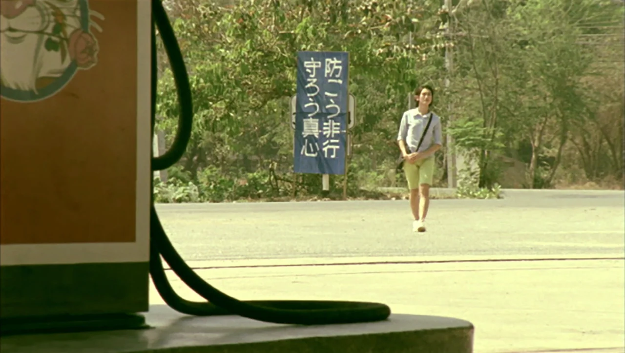 Wild Zero (1999) movie still: wide — A man walks past a blue Japanese text sign at a rural gas station. The foreground features the base and nozzle…